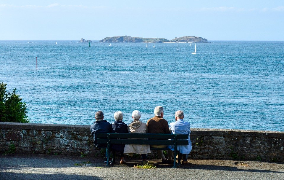 A group of old people sat on a bench looking out to sea.